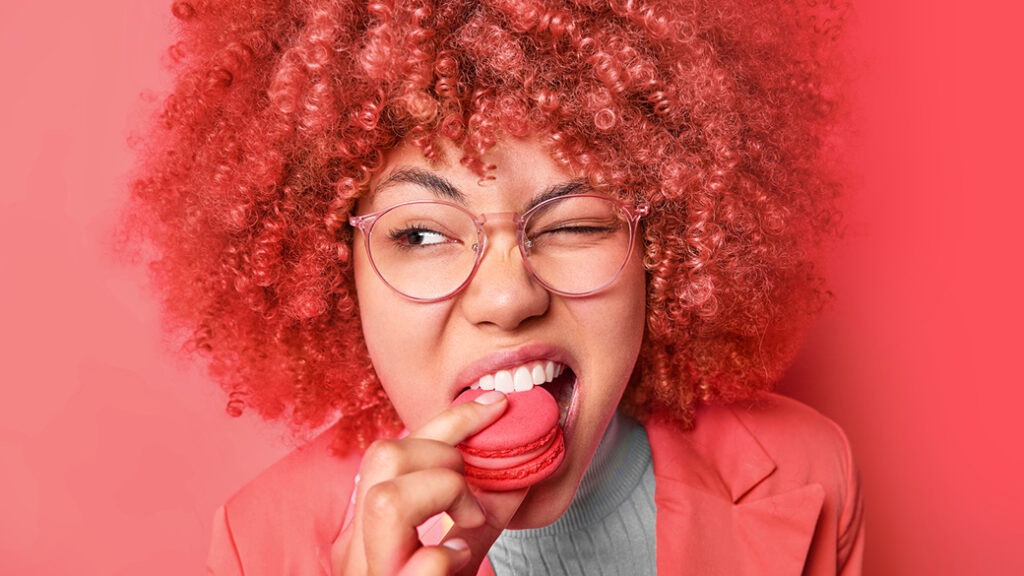 Ein Foto einer Frau mit roten Afrolocken die in ein rotes Macaron beißt.