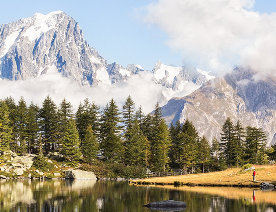 Landschaftsaufnahme eines Bergsees, man sieht eine in Rot gekleidete Person am Ufer stehen und große Berge im Hintergrund.