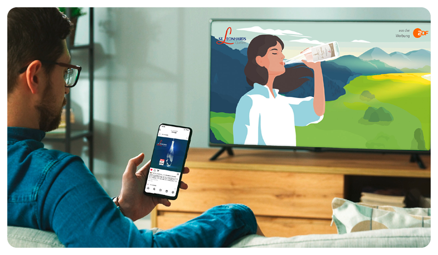 Ein Mann schaut auf ein Smartphone und einen Fernseher, die beide Werbung für St. Leonhardsquelle zeigen.