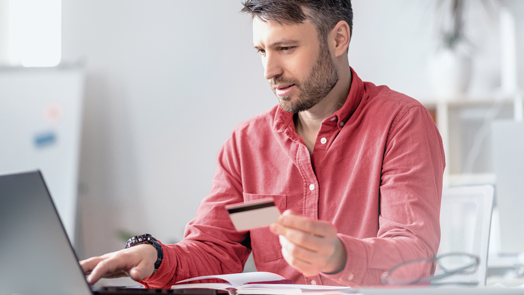 Foto eines Mannes, der eine Bankkarte in der Hand hält und gerade etwas in seinen Laptop tippt.