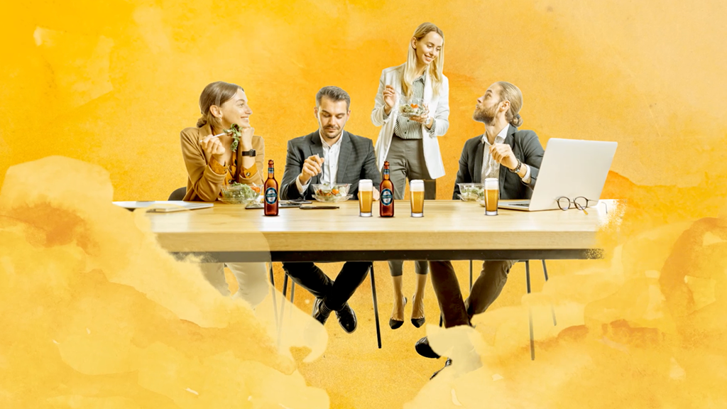 Foto einer Gruppe von Menschen, die an einem Tisch mit Getränken und Salaten sitzen und stehen, vor einem gelben Hintergrund.