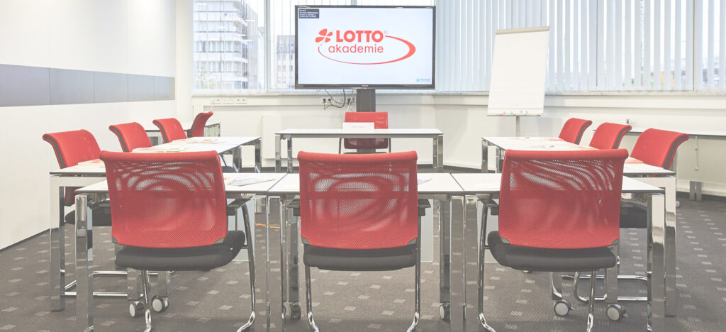 Moderner Seminarraum mit roten Stühlen, Tischen, einem Flipchart und einem Bildschirm mit dem Logo der LOTTO Akademie.
