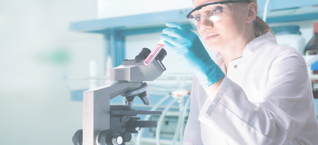 Eine Wissenschaftlerin in einem Labor, die eine Probe in einem Reagenzglas hält und ein Mikroskop verwendet.