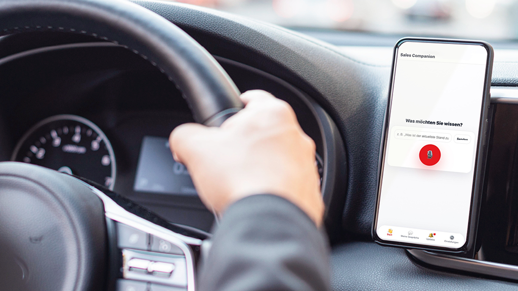 Hand am Lenkrad, Smartphone an Autoarmatur mit Sales Companion-App, Fokus auf Sprachsuche.