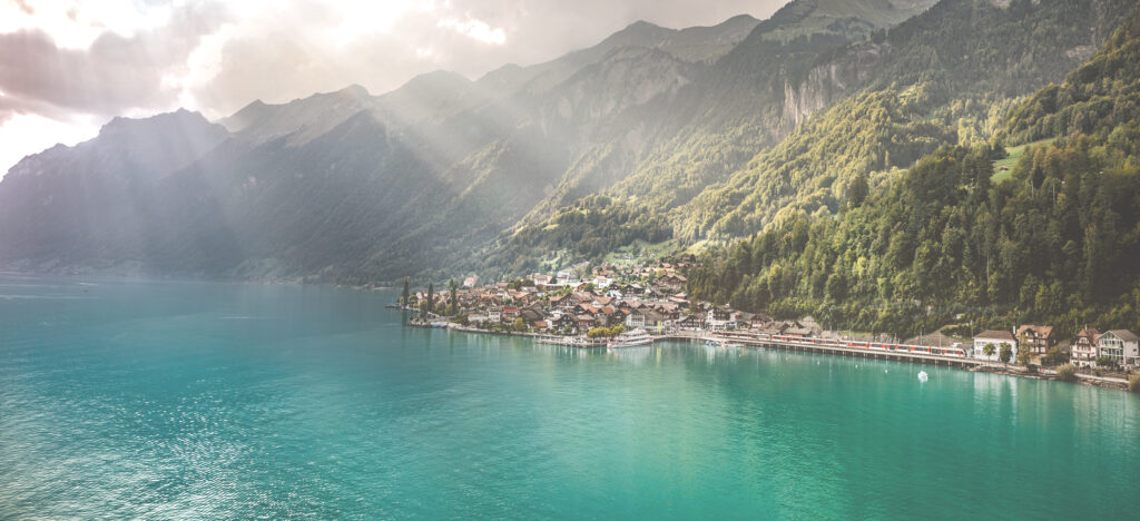 Landschaftsbild eines blauen Sees an dessen Ufer ein kleines Dorf steht, hinter dem Dorf beginnen die Berge.
