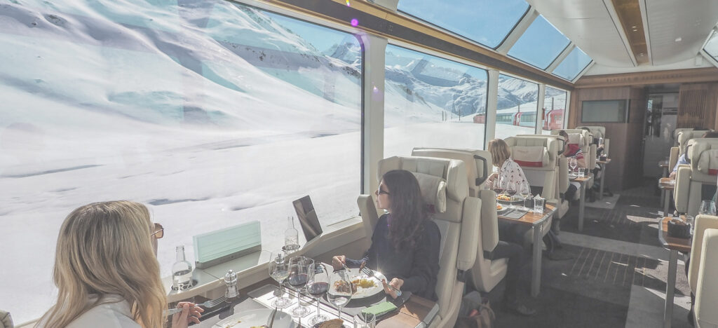 Foto aus dem inneren eines Panorama Zuges. Gäste sitzen in gemütlichen Zugsitzen und genießen ein Mittagessen.
