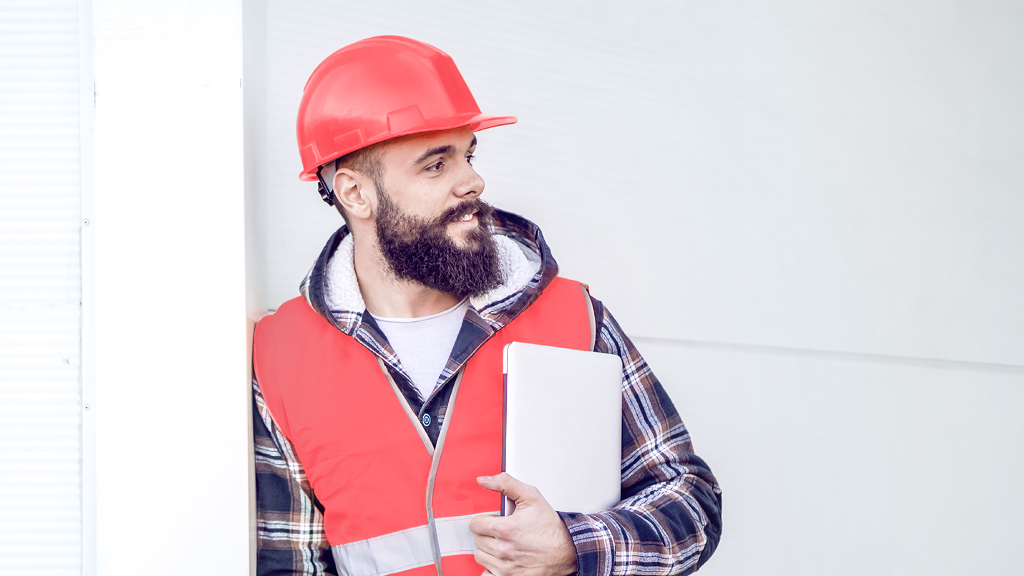Ein Arbeiter mit roter Warnweste und Helm steht an die Wand gelehnt und lächelt.